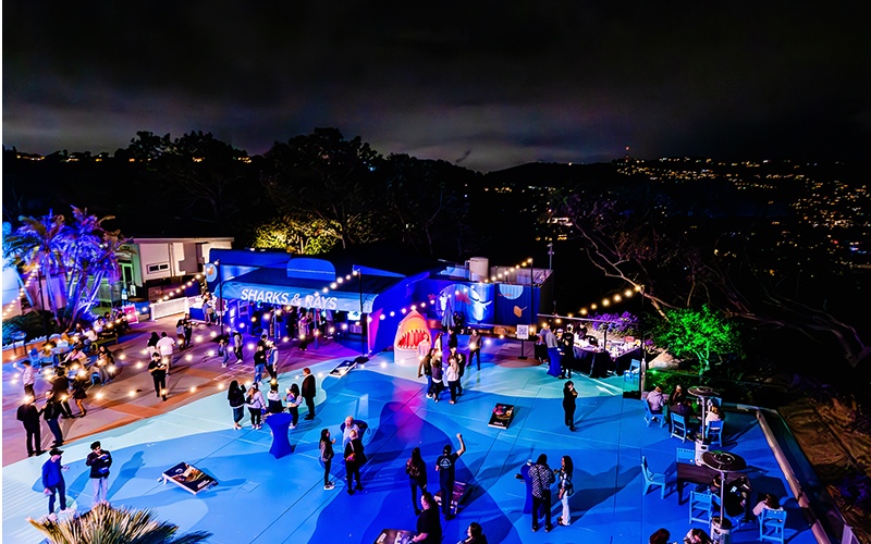 A nighttime view of Blue Beach illuminated by market lights, with people enjoying games like cornhole. The scene overlooks the ocean, creating a lively yet serene coastal ambiance.