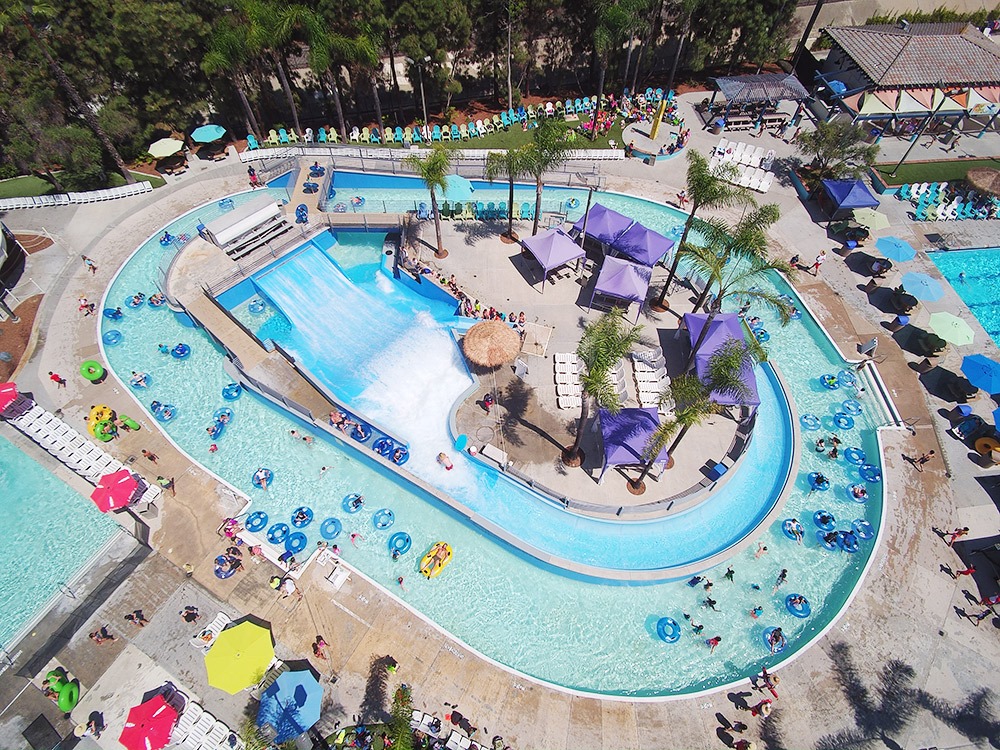 Aerial view of a water park featuring a lazy river, a simulated wave surfing area, and shaded lounge areas with purple cabanas and palm trees. People are floating in inner tubes, swimming, and relaxing on poolside chairs under colorful umbrellas. The park is surrounded by trees and includes several pool areas and shaded seating.