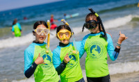 Three campers wearing Birch Aquarium green and blue rash guards and snorkel gear stand in front of the ocean