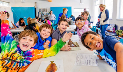 A group of cheerful students in a classroom, some wearing brightly colored shirts, engaging in an science project and looking at the camera with smiles. Teachers and other students are visible in the background.