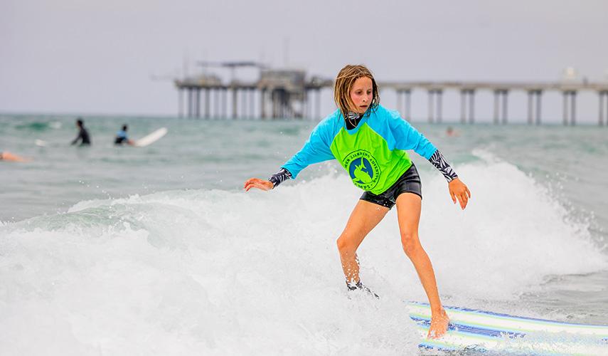 A student rides a surfboard on a small wave in front of Scripps Pier.