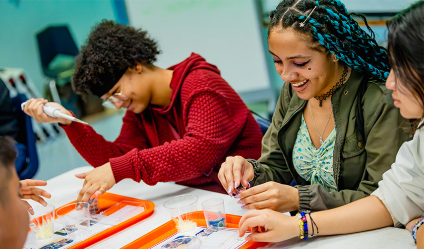 A group of students engages in a science experiment in a classroom, using test tubes and trays on a table. They are smiling and focused on their work.